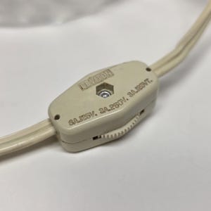 May include: A close-up of a cream-colored Leviton inline cord switch. The switch is rectangular with rounded edges and a small wheel for adjusting the light. The cord is a matching cream color.