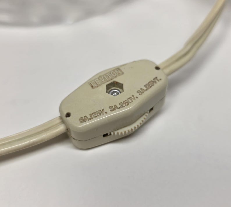 May include: A close-up of a cream-colored Leviton inline cord switch. The switch is rectangular with rounded edges and a small wheel for adjusting the light. The cord is a matching cream color.