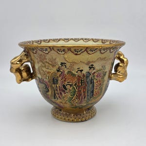 Peut inclure: Un bol en céramique avec une finition dorée et deux anses ornées de couleur or. L'extérieur est décoré d'une scène détaillée représentant des personnages en vêtements traditionnels parmi des arbres et des fleurs, avec un bord et une base dorés.
