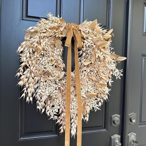 May include: A circular wreath composed of dried beige and tan botanical elements, including various grasses, leaves, and small sprigs. A long, smooth, light brown velvet ribbon is tied at the top center, with two long tails hanging down the middle.