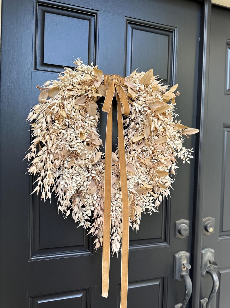 May include: A circular wreath composed of dried beige and tan botanical elements, including various grasses, leaves, and small sprigs. A long, smooth, light brown velvet ribbon is tied at the top center, with two long tails hanging down the middle.