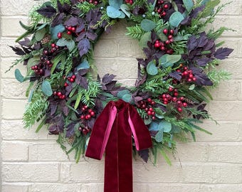 May include: A festive wreath with a mix of greenery, deep purple leaves, and bright red berries. A large, burgundy velvet bow hangs at the bottom. The wreath is displayed against a white brick wall, perfect for holiday decor.