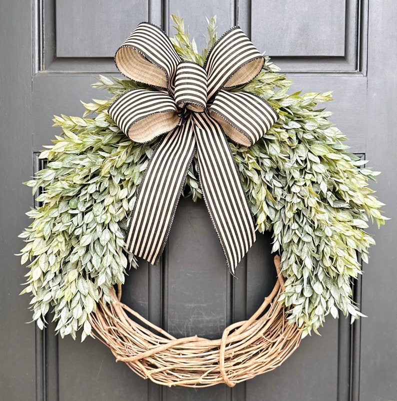 May include: A wreath made of brown vines with a black and white striped bow. The wreath is decorated with green leaves and is hanging on a gray door.