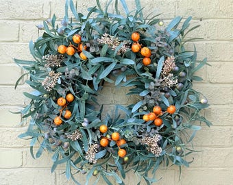 May include: A circular wreath featuring sage green elongated leaves, clusters of bright orange berries, and small sprigs of light brown textured filler foliage. The arrangement is dense and layered, creating a full, rounded appearance with varied botanical elements throughout the design.