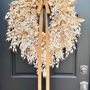 May include: A heart-shaped wreath constructed from dried beige and tan foliage, including various leaves and small seed pods. A long, smooth, golden-brown velvet ribbon is tied at the top center, with two long tails hanging down the middle of the wreath.