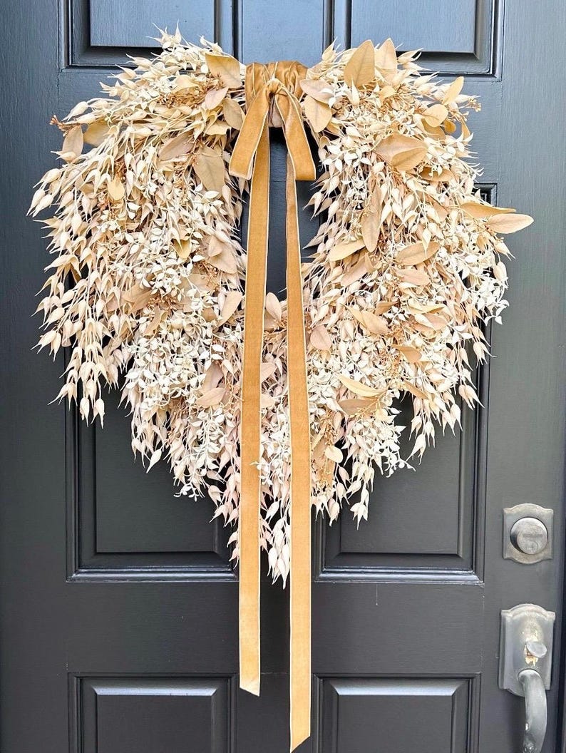 May include: A heart-shaped wreath constructed from dried beige and tan foliage, including various leaves and small seed pods. A long, smooth, golden-brown velvet ribbon is tied at the top center, with two long tails hanging down the middle of the wreath.