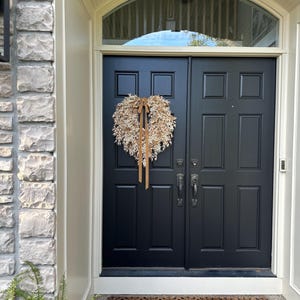 May include: A heart-shaped wreath made of dried beige foliage and grasses, featuring a long, thin, light brown ribbon tied at the top center, hanging on a solid black double door with rectangular panels and dark metal hardware.
