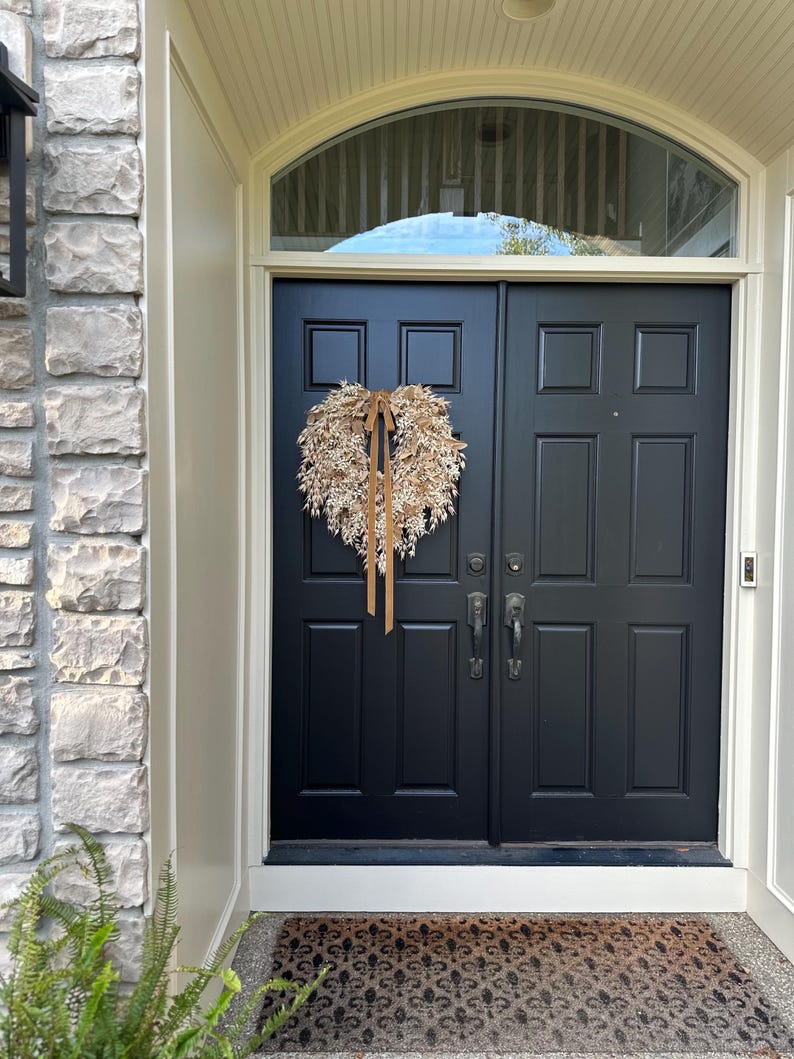 May include: A heart-shaped wreath made of dried beige foliage and grasses, featuring a long, thin, light brown ribbon tied at the top center, hanging on a solid black double door with rectangular panels and dark metal hardware.