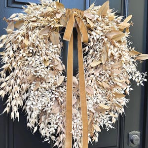 May include: A circular wreath composed of dried, light beige and tan botanical elements, including small leaves, seed pods, and delicate sprigs. A long, smooth, golden-brown velvet ribbon is tied at the top, with two long tails hanging down the center.