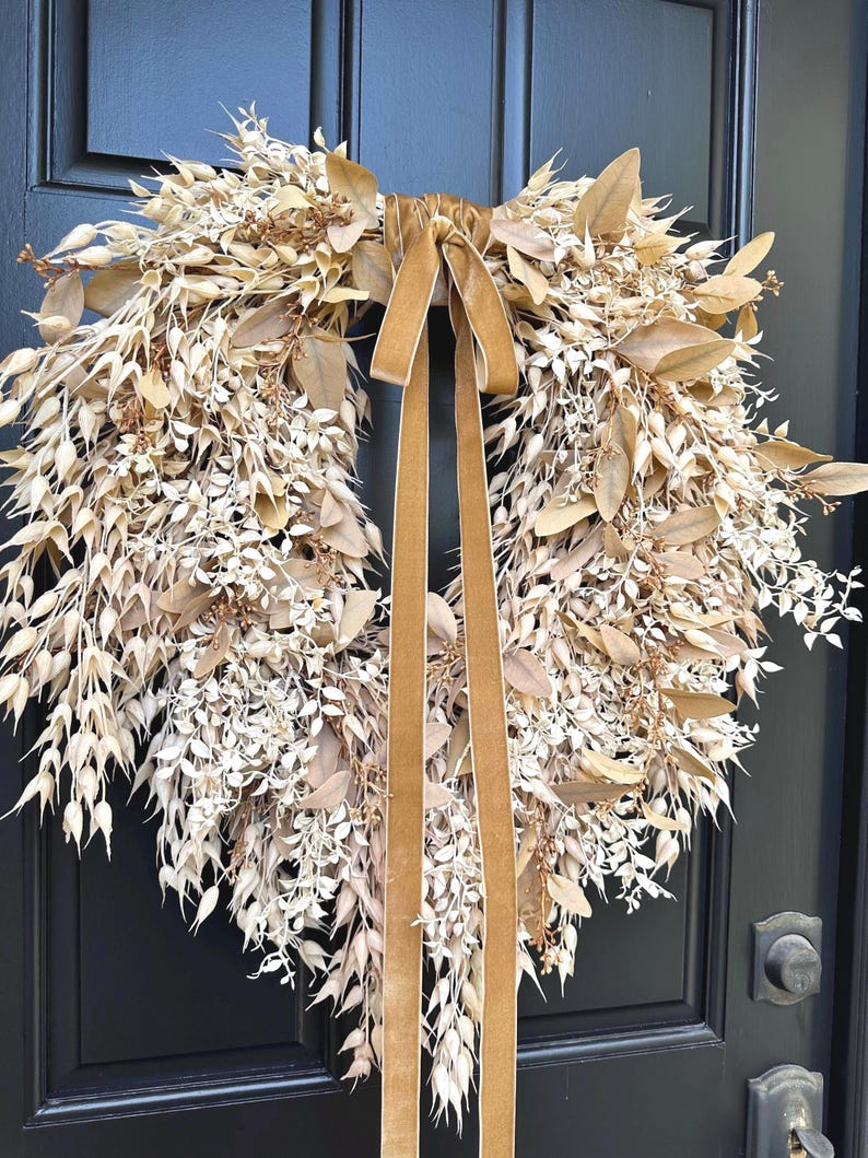 May include: A circular wreath composed of dried, light beige and tan botanical elements, including small leaves, seed pods, and delicate sprigs. A long, smooth, golden-brown velvet ribbon is tied at the top, with two long tails hanging down the center.