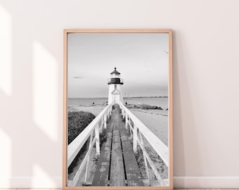 May include: Black and white framed print of a lighthouse at the end of a wooden pier. The pier has white railings and leads to the lighthouse. The print is in a light brown frame and is displayed on a light wood floor.