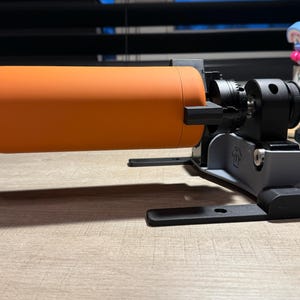 May include: A 3D printed device with a rotating platform holding an orange water bottle. The device has a black base with adjustable legs and a gray arm with a knob.