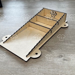 May include: A wooden organiser with three compartments, designed to hold small items. The organiser is made of light brown wood and has a black outline. It has a cutout design on the top with a stylized image of a house.