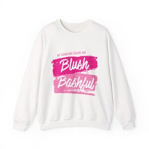 May include: White sweatshirt featuring the text "MY SIGNATURE COLORS ARE" above the words "Blush AND Bashful" in pink and light pink brushstrokes. The sweatshirt has a crew neck and long sleeves.