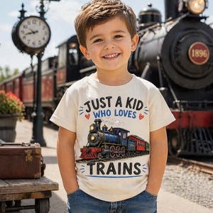 May include: A young child wearing a cream-colored t-shirt with the text "JUST A KID WHO LOVES TRAINS" and a graphic of a blue, red, and black train. The background includes a vintage train and a train station sign.