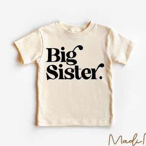 May include: A cream colored t-shirt with black text that reads "Big Sister."