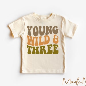 May include: A cream colored t-shirt with the text "YOUNG WILD & THREE" in brown, orange, and green lettering. The shirt is a toddler size and has short sleeves.