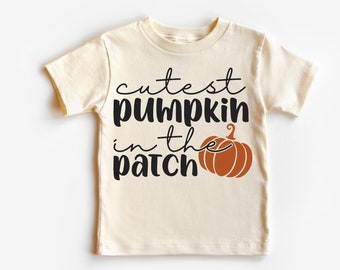 May include: Cream-colored t-shirt with the phrase "cutest pumpkin in the patch" in black script and a brown pumpkin graphic. The shirt is a short-sleeved crew neck, perfect for fall. The brand name "Madi Moosh" is printed at the bottom.