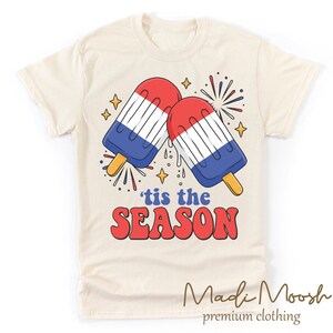 May include: A beige t-shirt with a red, white, and blue popsicle graphic and the text 'tis the SEASON'.