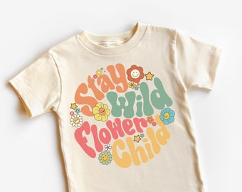 May include: Cream-colored t-shirt with the text "Stay Wild Flower Child" in a colorful, retro font. The design includes various smiling flowers and stars. The brand name "Madi Moosh" is printed at the bottom.