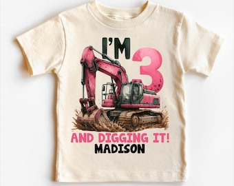 May include: Cream-colored toddler t-shirt featuring a pink excavator graphic with the number 3. Text on the shirt reads "I'M 3 AND DIGGING IT! MADISON". The excavator is depicted in a construction scene.