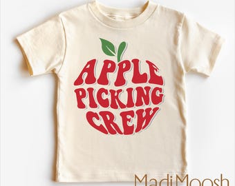 May include: Cream-colored t-shirt with a red apple graphic and the words "APPLE PICKING CREW." The apple has green leaves. The brand name "Madi Moosh" is printed at the bottom. Made of soft material.