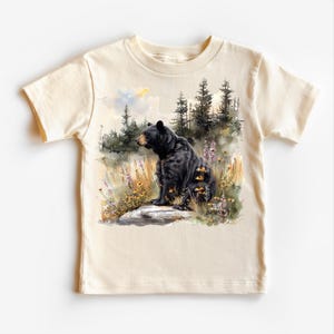 May include: A cream-colored t-shirt featuring a watercolor illustration of a black bear sitting on a rock in a field of wildflowers. The bear is facing right, with a forest backdrop. The shirt has short sleeves and a crew neck.