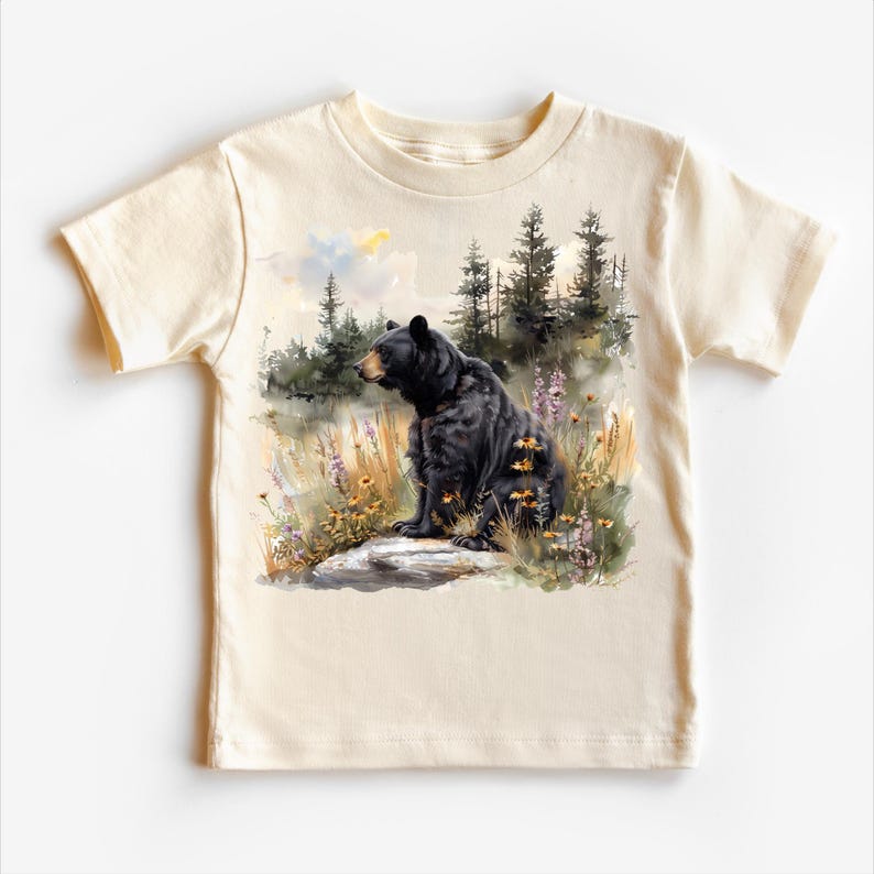 May include: A cream-colored t-shirt featuring a watercolor illustration of a black bear sitting on a rock in a field of wildflowers. The bear is facing right, with a forest backdrop. The shirt has short sleeves and a crew neck.