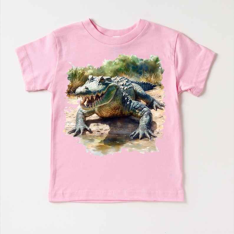 K&ouml;nnte beinhalten: Rosafarbenes T-Shirt f&uuml;r Kleinkinder mit einer Aquarell-Illustration eines Alligators in einer Sumpflandschaft.