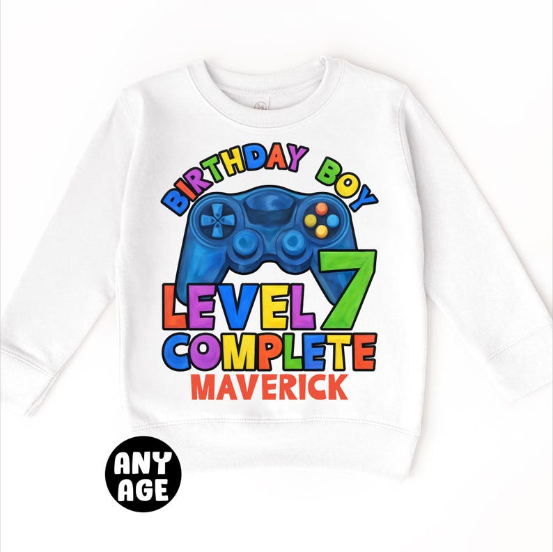 May include: White long-sleeve sweatshirt with a colorful graphic of a video game controller and the text "Birthday Boy Level 7 Complete Maverick".