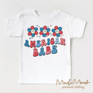 May include: White t-shirt featuring the text "AMERICAN BABE" in red and blue bubble letters. Above the text are three smiling flower graphics in red and blue. The shirt has a retro, patriotic design.