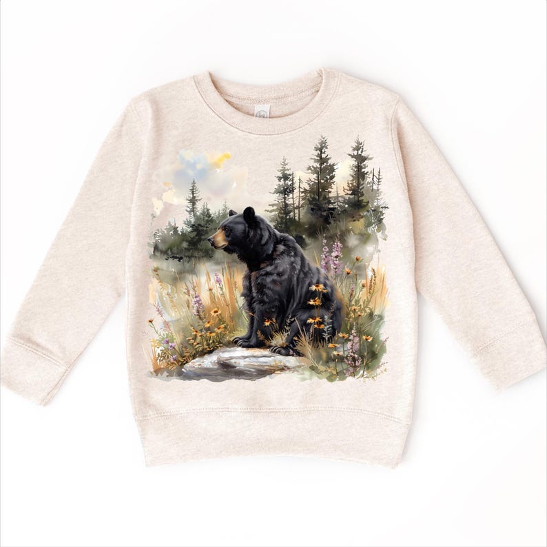 May include: A beige sweatshirt featuring a watercolor illustration of a black bear sitting on a rock in a field of wildflowers. The bear is facing left, with a forest scene in the background. The sweatshirt has long sleeves and a crew neck.