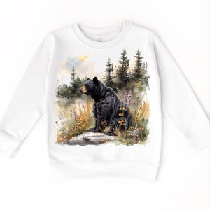 May include: White sweatshirt featuring a detailed watercolor painting of a black bear sitting on a rock in a forest setting. The bear is surrounded by wildflowers and tall grass, with a backdrop of pine trees and a soft, cloudy sky.