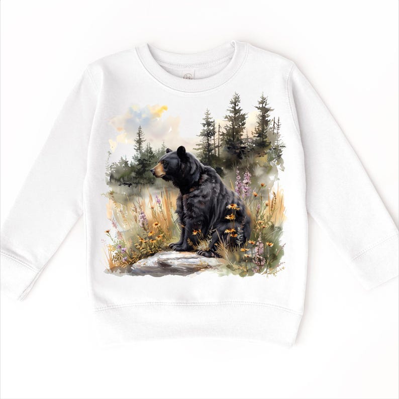 May include: White sweatshirt featuring a detailed watercolor painting of a black bear sitting on a rock in a forest setting. The bear is surrounded by wildflowers and tall grass, with a backdrop of pine trees and a soft, cloudy sky.