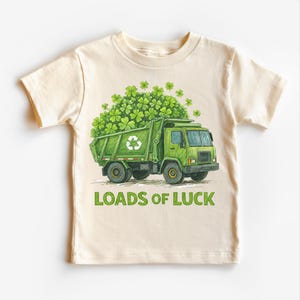 May include: Cream-colored t-shirt featuring a green garbage truck overflowing with green clovers. The truck has a recycling symbol on the side. The words "LOADS OF LUCK" are printed in green below the truck.