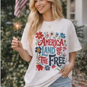 May include: Cream-colored t-shirt with the text "AMERICA LAND OF THE FREE" in red, blue, and teal, surrounded by floral designs. The person is holding a lit sparkler. An American flag is in the background.