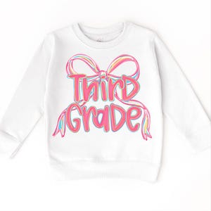 May include: White long-sleeved sweatshirt featuring a colorful bow design with the words "Third Grade" in pink and blue lettering. The sweatshirt is a comfortable, casual garment for children.