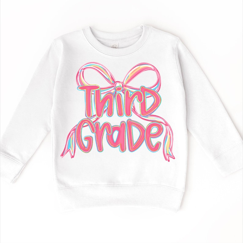 May include: White long-sleeved sweatshirt featuring a colorful bow design with the words "Third Grade" in pink and blue lettering. The sweatshirt is a comfortable, casual garment for children.