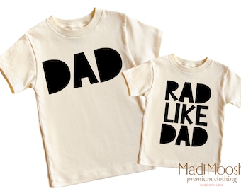 May include: Two cream colored t-shirts with black text. The larger shirt says "DAD" and the smaller shirt says "RAD LIKE DAD".