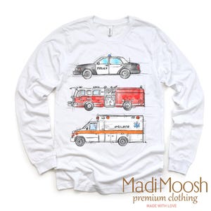 May include: White long-sleeve t-shirt with a graphic print of a police car, fire truck, and ambulance. The vehicles are drawn in a cartoon style with watercolor accents. The text "Madi Moosh premium clothing" is printed on the bottom of the shirt.