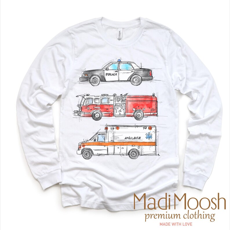 May include: White long-sleeve t-shirt with a graphic print of a police car, fire truck, and ambulance. The vehicles are drawn in a cartoon style with watercolor accents. The text "Madi Moosh premium clothing" is printed on the bottom of the shirt.