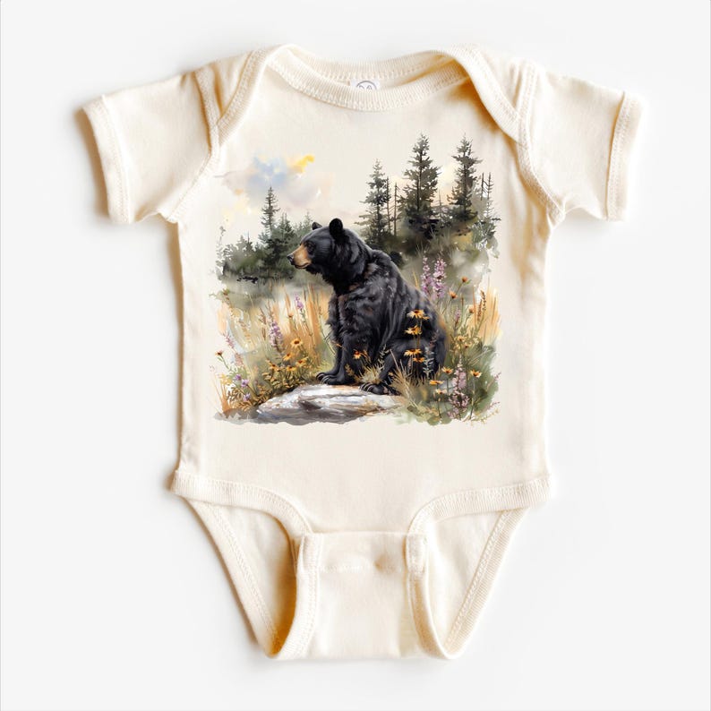 May include: Cream-colored baby onesie featuring a watercolor illustration of a black bear sitting on a rock. The bear is surrounded by wildflowers and trees, with a soft, natural background. The design is centered on the front of the garment.