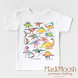 May include: White t-shirt with a colorful dinosaur print. The dinosaurs are all different colors and have names written underneath them. The shirt has the text "Madi Moosh premium clothing MADE WITH LOVE" at the bottom.
