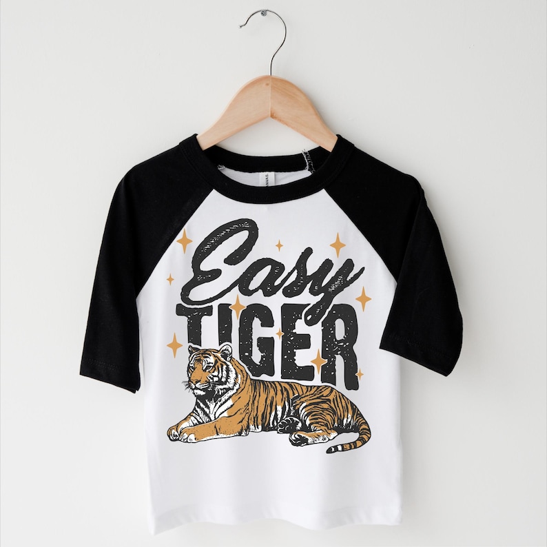 Op de afbeelding: Wit en zwart baseball shirt met een zwart en oranje tijger motief en de tekst "Easy Tiger" in zwart met gouden sterren.