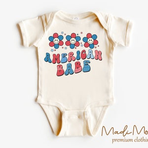 May include: Cream-colored baby bodysuit with the text "AMERICAN BABE" in red, white, and blue, along with smiling flower graphics and stars. The design has a retro, vintage style, perfect for patriotic celebrations.