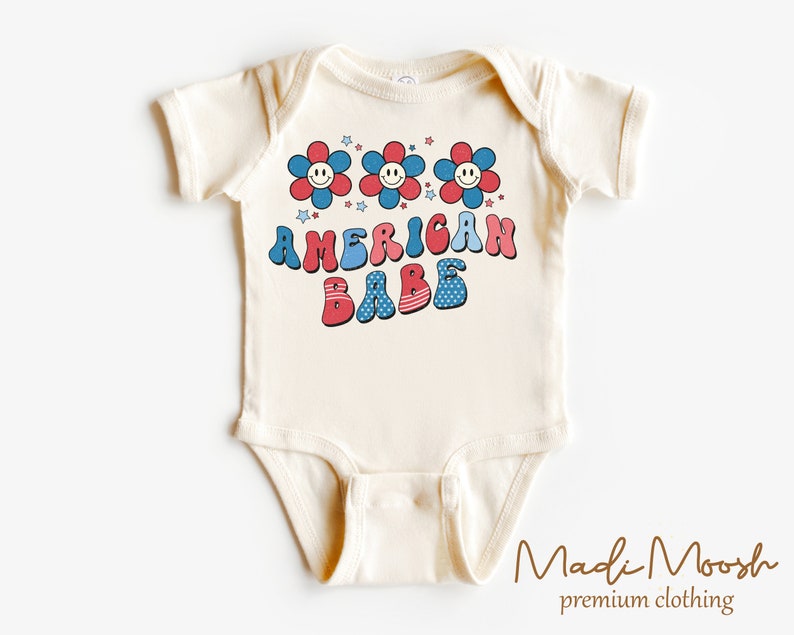 May include: Cream-colored baby bodysuit with the text "AMERICAN BABE" in red, white, and blue, along with smiling flower graphics and stars. The design has a retro, vintage style, perfect for patriotic celebrations.