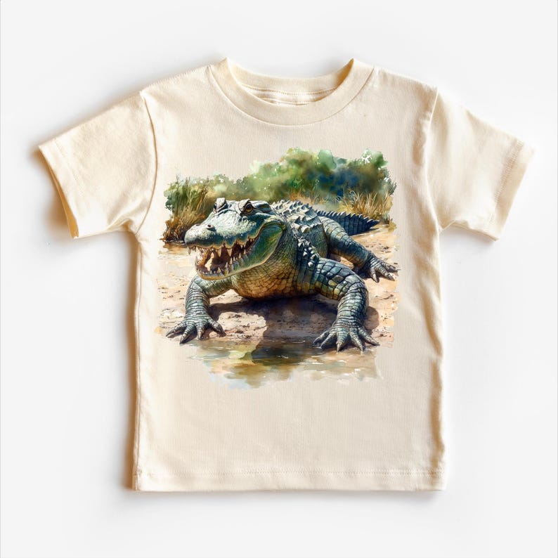 K&ouml;nnte beinhalten: Ein beiges T-Shirt mit einer Aquarell-Illustration eines Alligators in einer sumpfigen Umgebung.