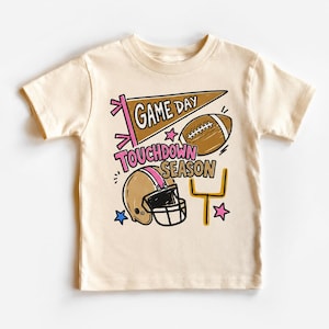 May include: A cream-colored short-sleeve t-shirt featuring a graphic print with a brown pennant that says GAME DAY, a brown football, a tan football helmet, a yellow goal post, and the text TOUCHDOWN SEASON in pink and brown lettering.
