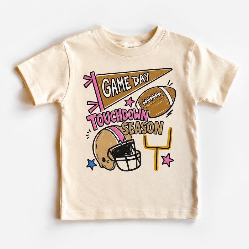 May include: A cream-colored short-sleeve t-shirt featuring a graphic print with a brown pennant that says GAME DAY, a brown football, a tan football helmet, a yellow goal post, and the text TOUCHDOWN SEASON in pink and brown lettering.