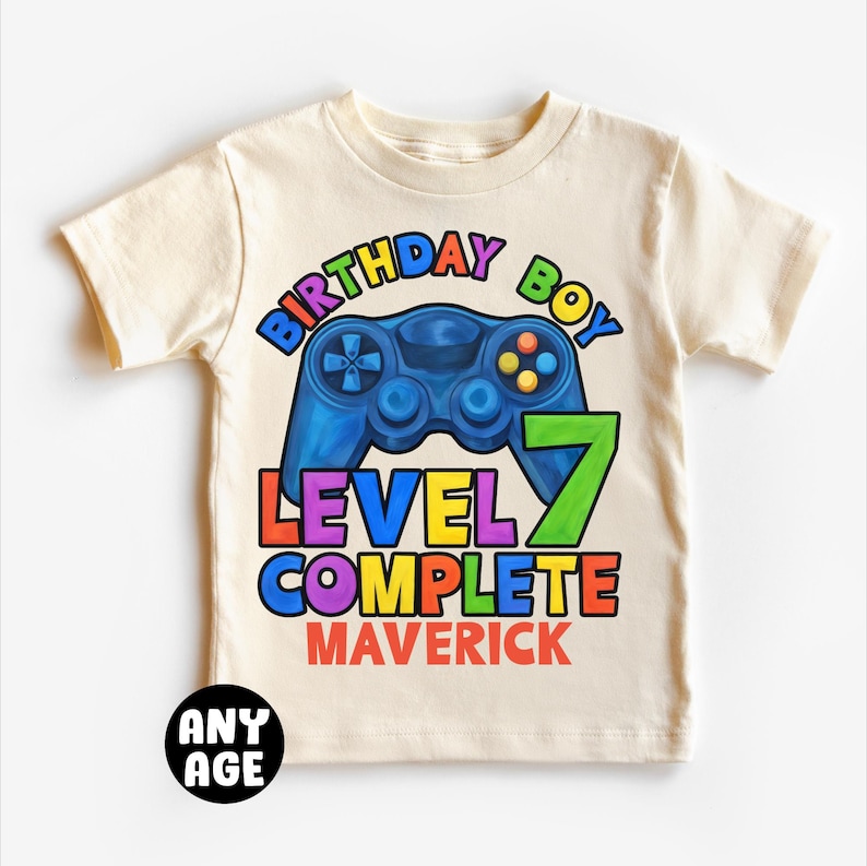 May include: A beige t-shirt with a colorful graphic of a video game controller and the text "Birthday Boy Level 7 Complete Maverick".